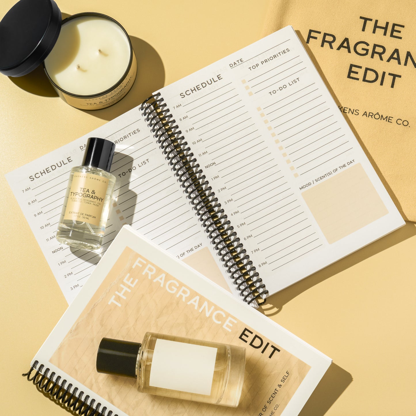 Two bottles of 'The Fragrance Edit' on a planner with a candle and notebook in the background.