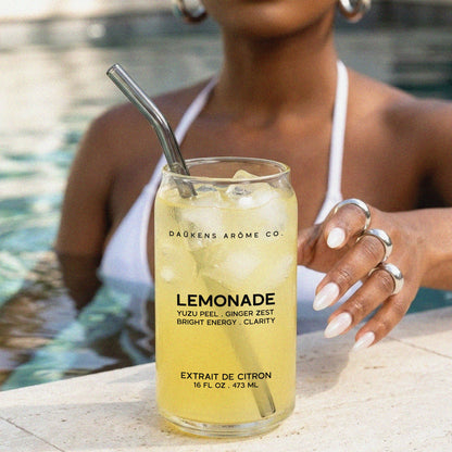 Person holding a glass of lemonade with a straw by a pool, featuring 'Dékens Arome Co.' branding.