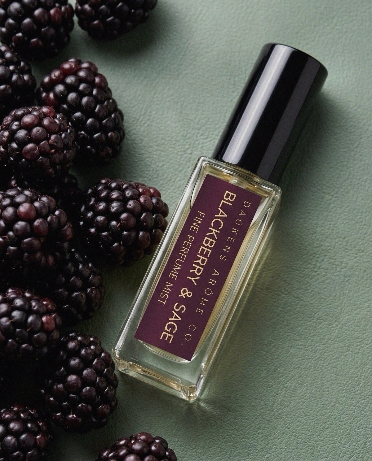 Blackberry & Sage Fine Perfume Mist bottle beside blackberries on a green surface.