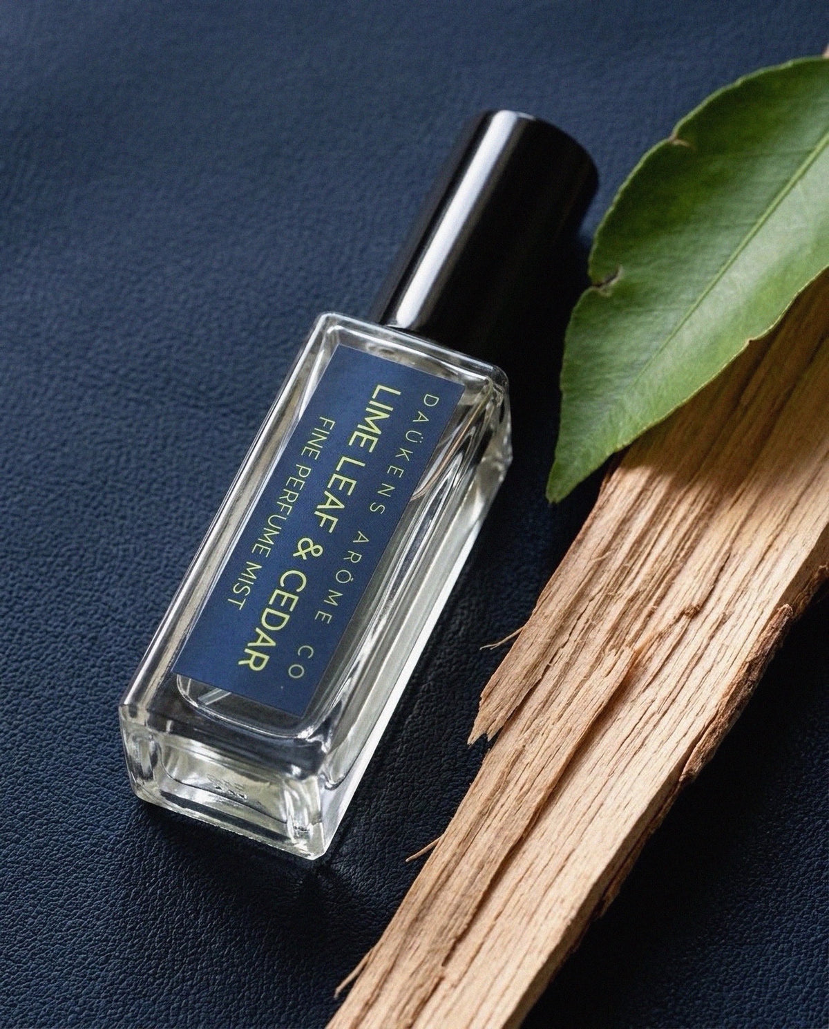 Lime Leaf & Cedar Fine Perfume Mist bottle beside a green leaf and wood on a blue surface.