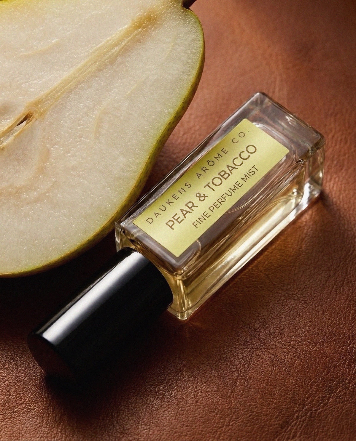 Pear & Tobacco Fine Perfume Mist bottle beside a cut pear on a brown surface.